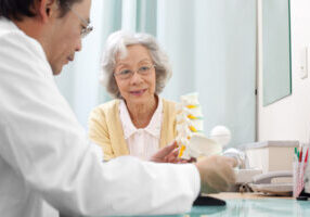 Senior woman visiting the doctor Senior woman visiting the doctor in his office. Doctor explaining spine problem to her using a bone model.