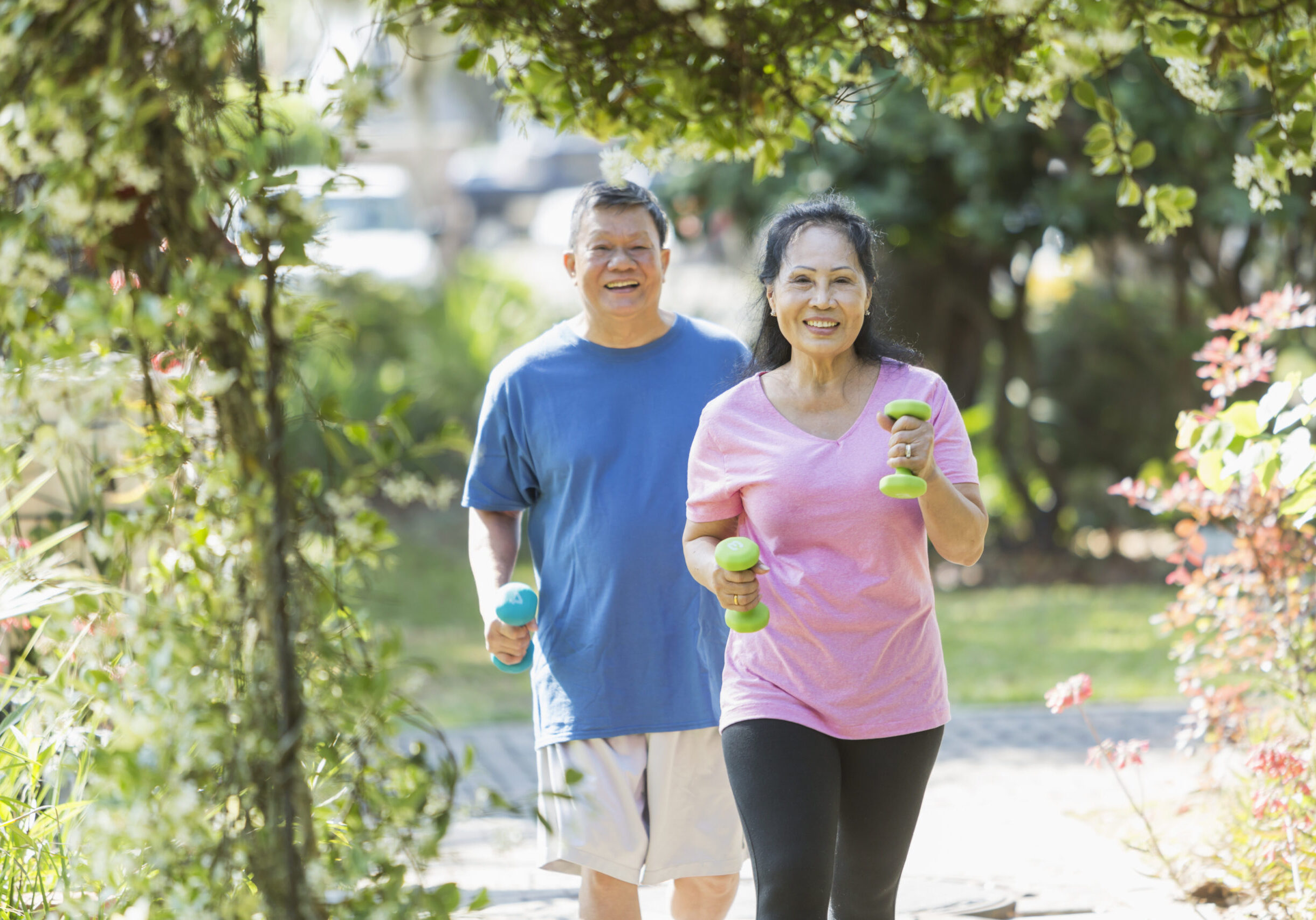A senior Asian couple in the park on a sunny day, exercising together. They are powerwalking or jogging with dumbbells in their hands.